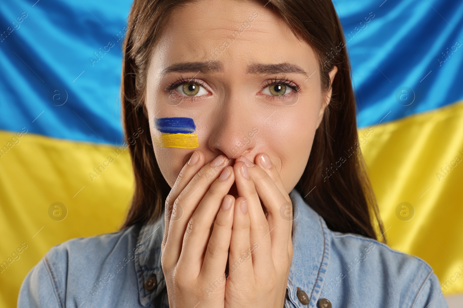 Photo of Sad young woman with clasped hands near Ukrainian flag, closeup