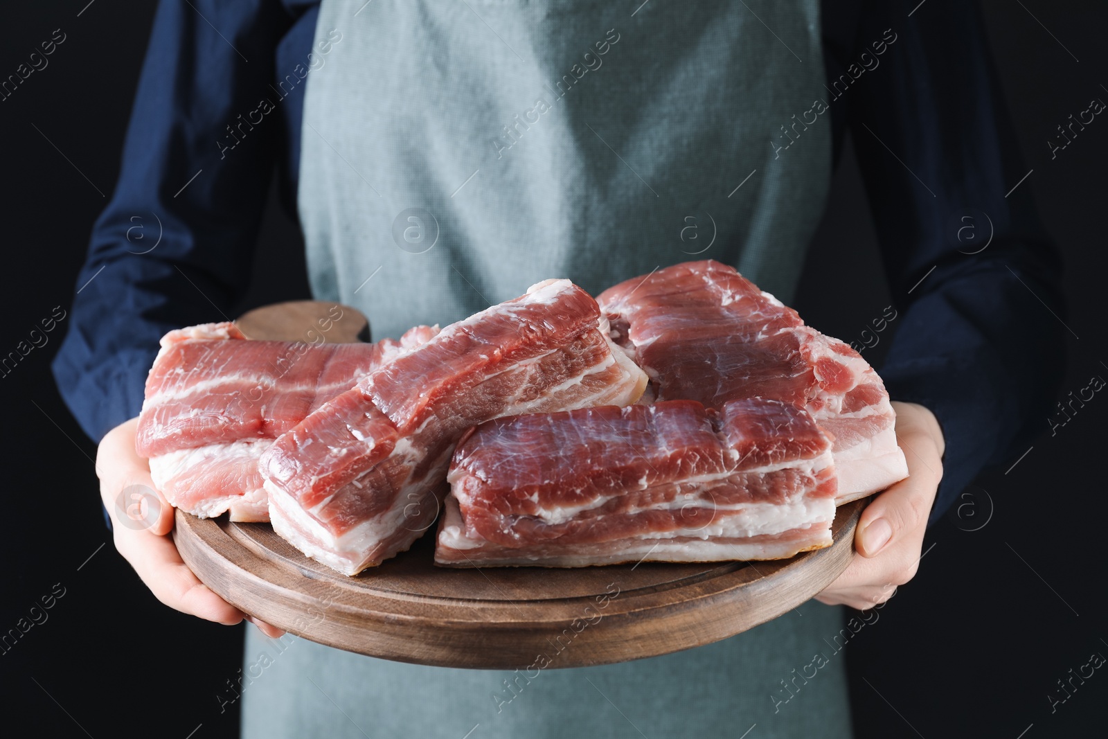 Photo of Woman holding wooden board with pieces of raw pork belly on black background, closeup