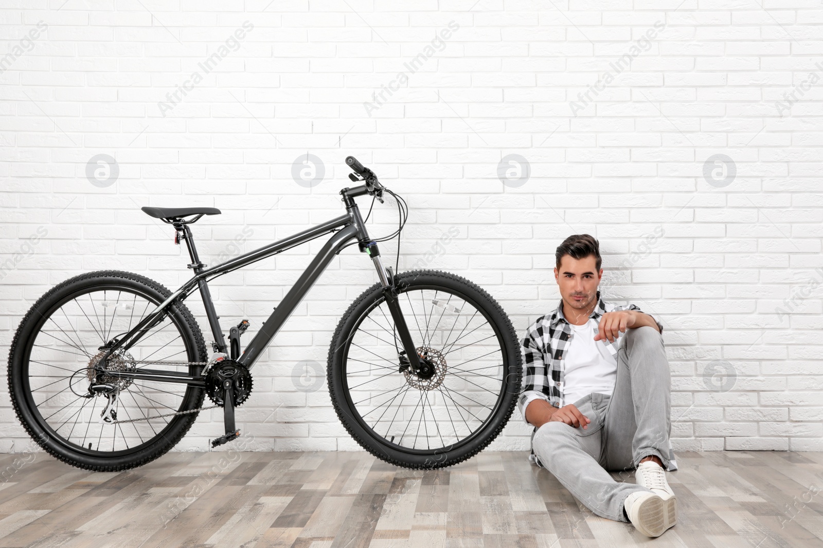 Photo of Handsome young man with modern bicycle near white brick wall indoors