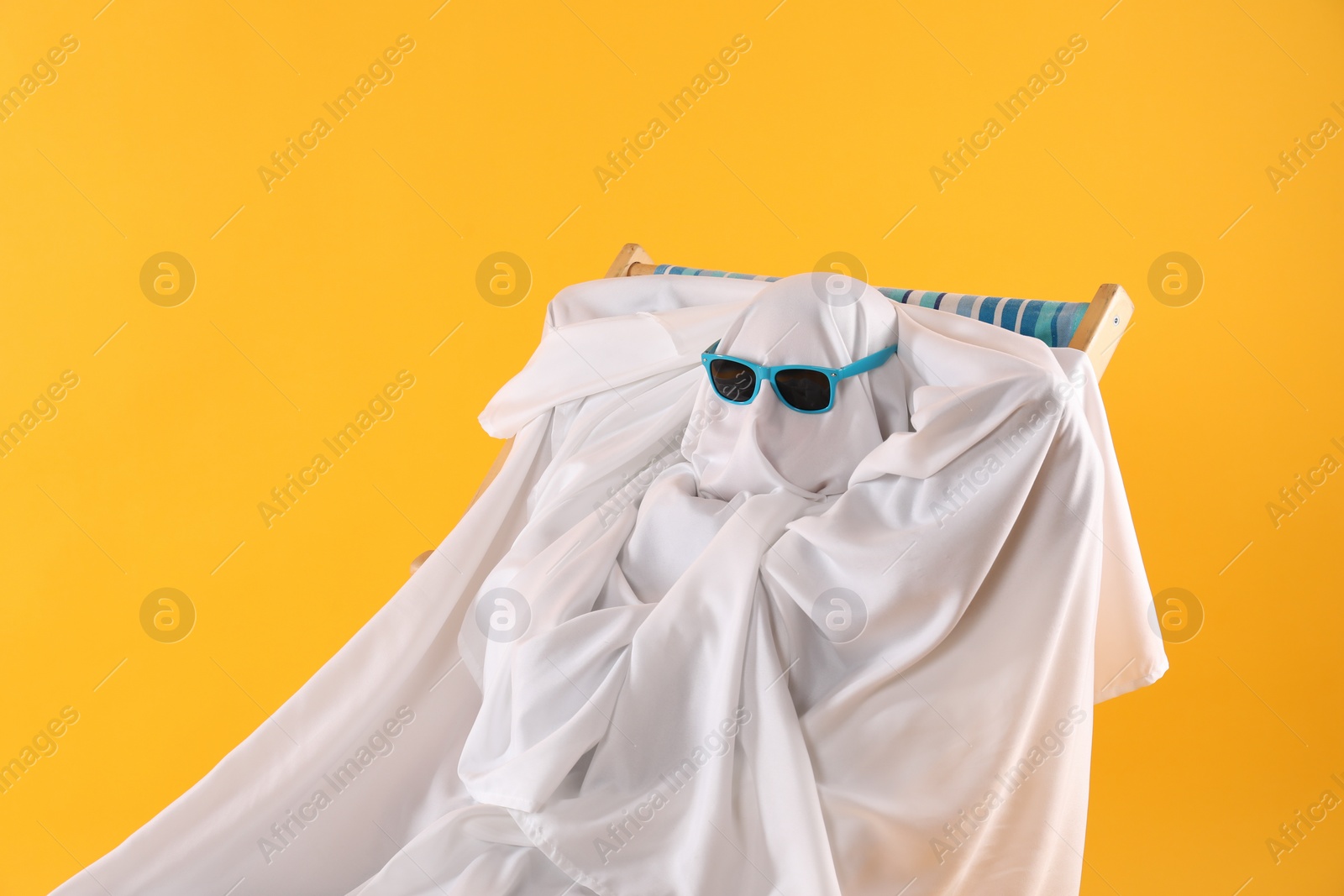 Photo of Person in ghost costume and sunglasses relaxing on deckchair against yellow background