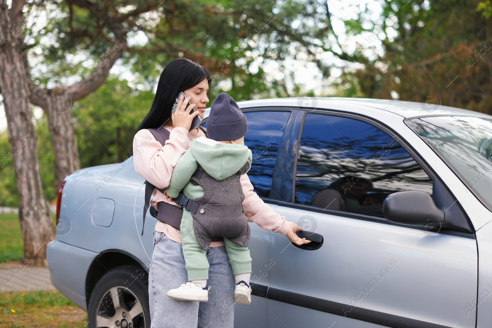 Photo of Mother holding her child in sling (baby carrier) while talking on smartphone near car outdoors