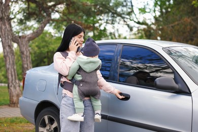 Mother holding her child in sling (baby carrier) while talking on smartphone near car outdoors