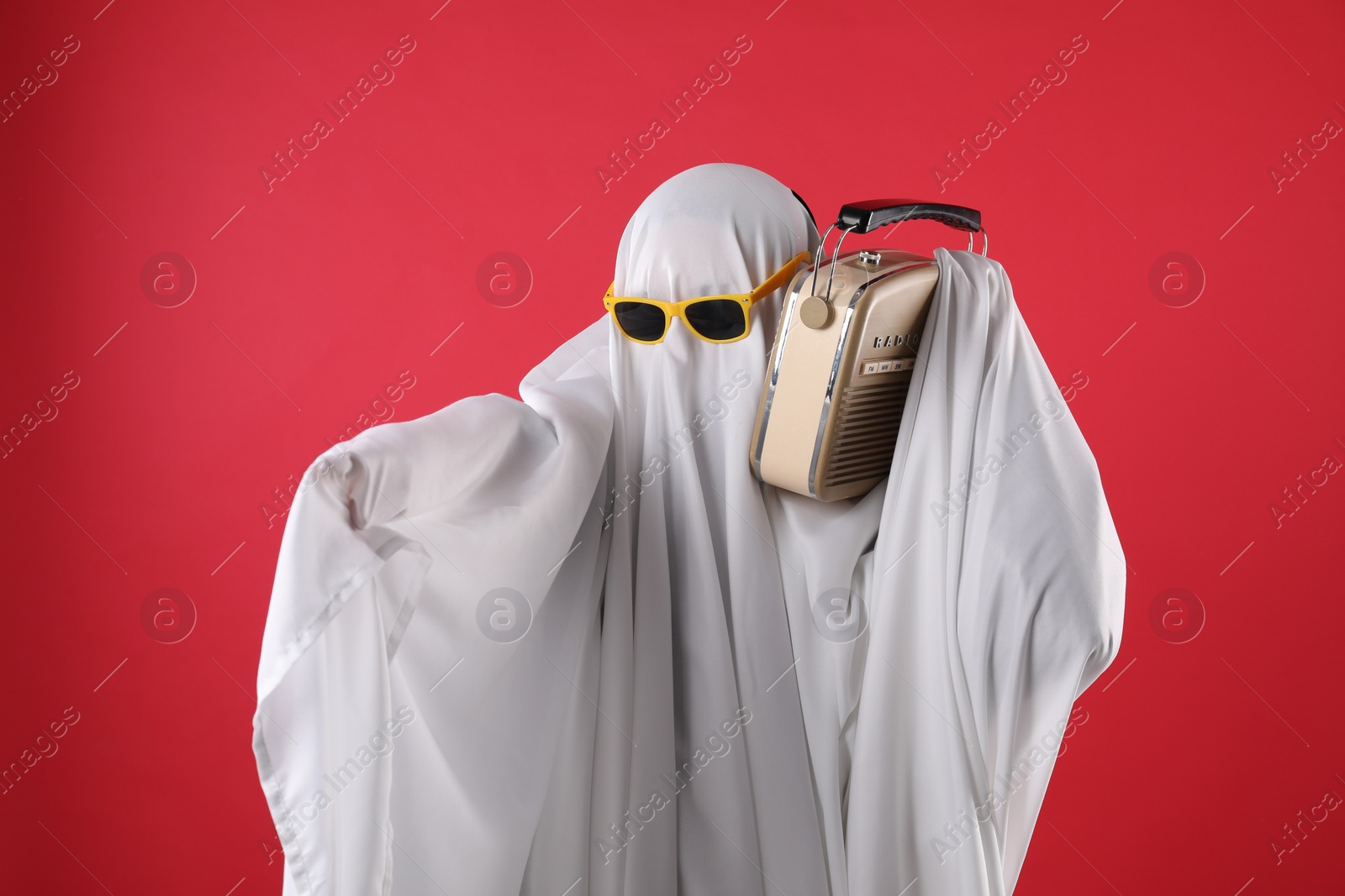 Photo of Person in ghost costume and sunglasses using retro radio receiver on red background