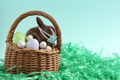 Photo of Chocolate bunny with protective mask and eggs in basket on light blue background, space for text. Easter holiday during COVID-19 quarantine