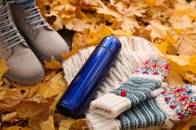 Photo of Woman standing near blue thermos, knitted scarf and mittens on fallen maple leaves outdoors, closeup