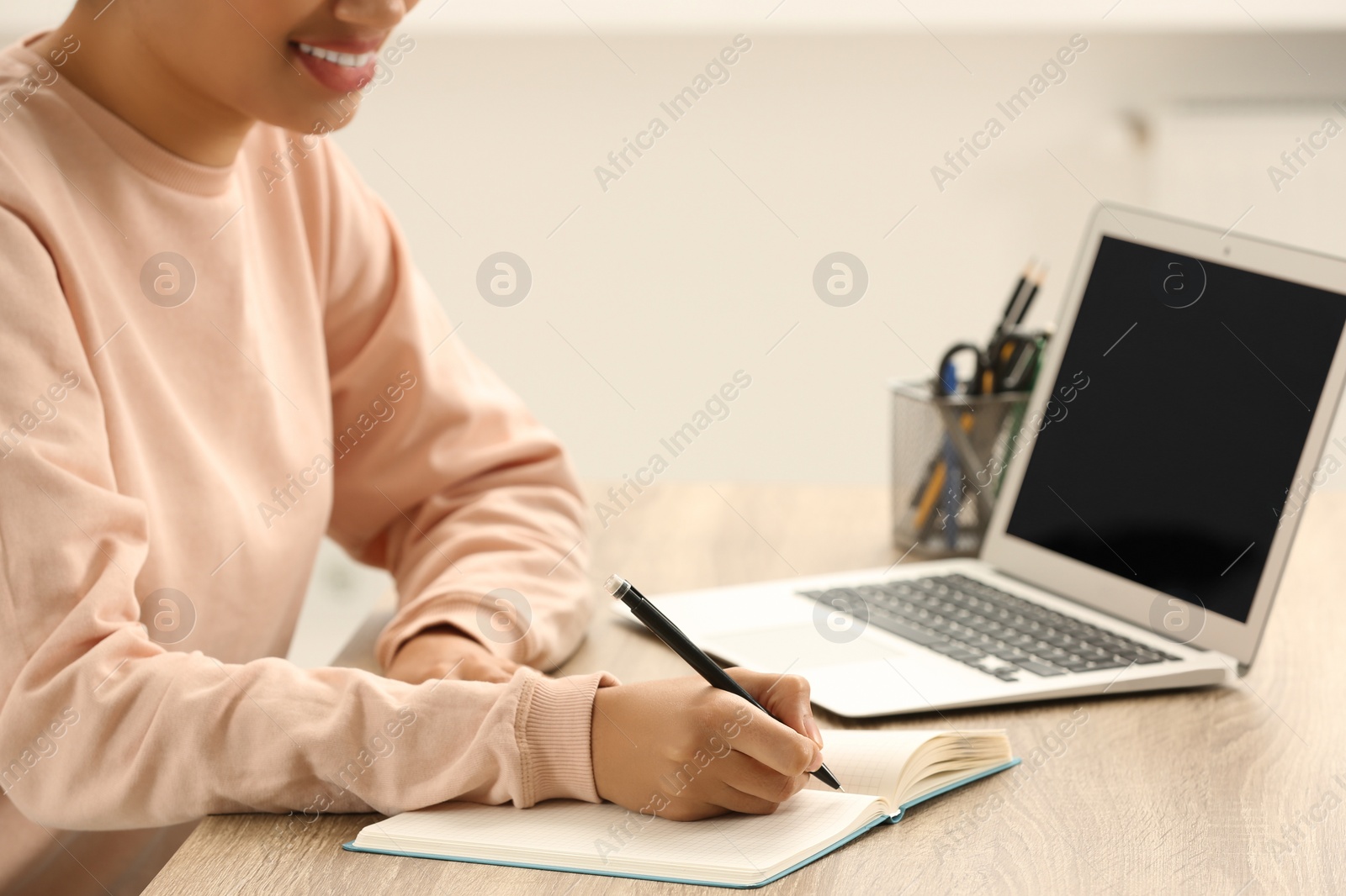 Photo of African American woman writing in notepad near laptop at wooden table indoors, closeup