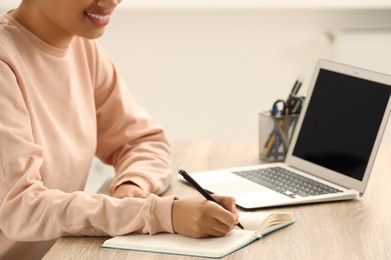 Photo of African American woman writing in notepad near laptop at wooden table indoors, closeup