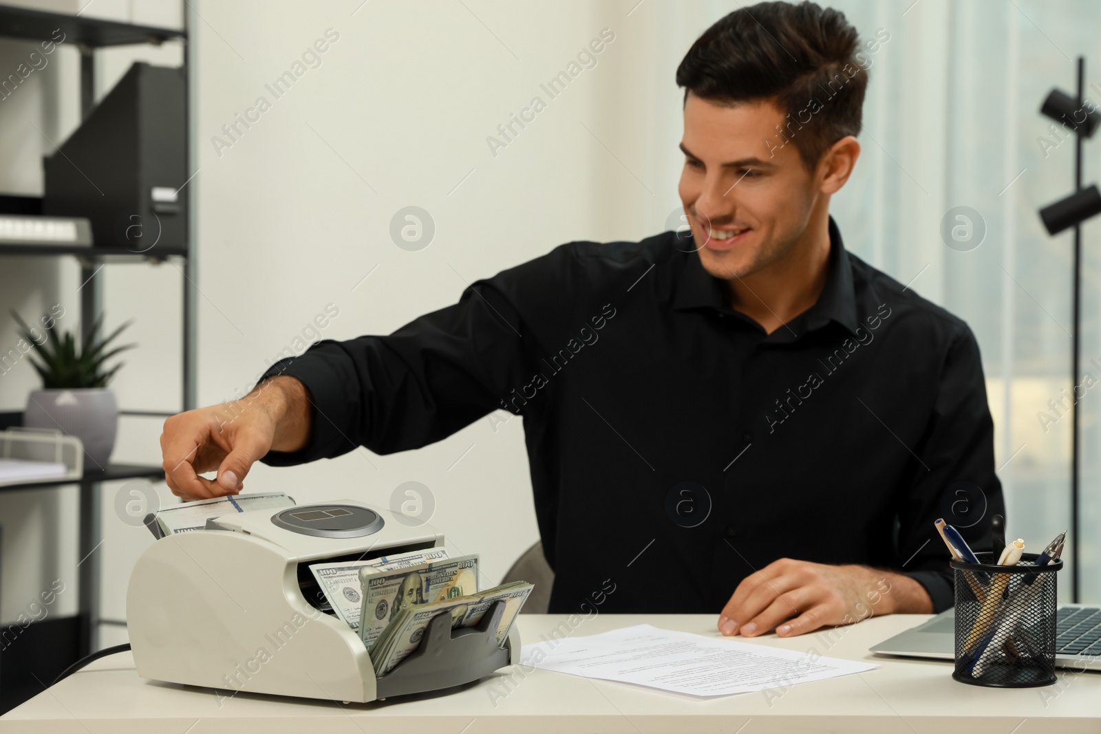 Photo of Man putting money into banknote counter at white table indoors