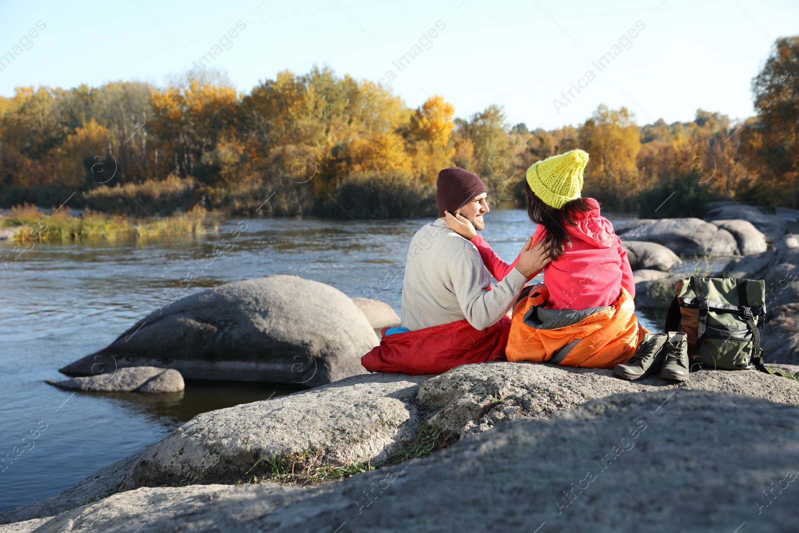 Photo of Couple of campers in sleeping bags sitting on rock near pond. Space for text