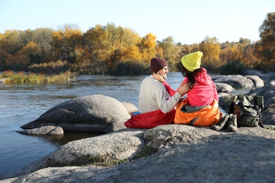 Photo of Couple of campers in sleeping bags sitting on rock near pond. Space for text