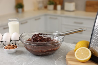 Metal whisk, chocolate cream in bowl and different products on gray marble table indoors