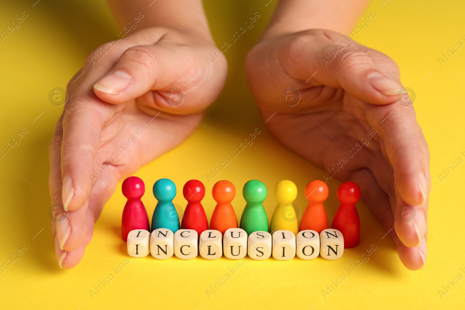 Photo of Woman protecting colorful pawns and wooden cubes with word Inclusion on yellow background, closeup