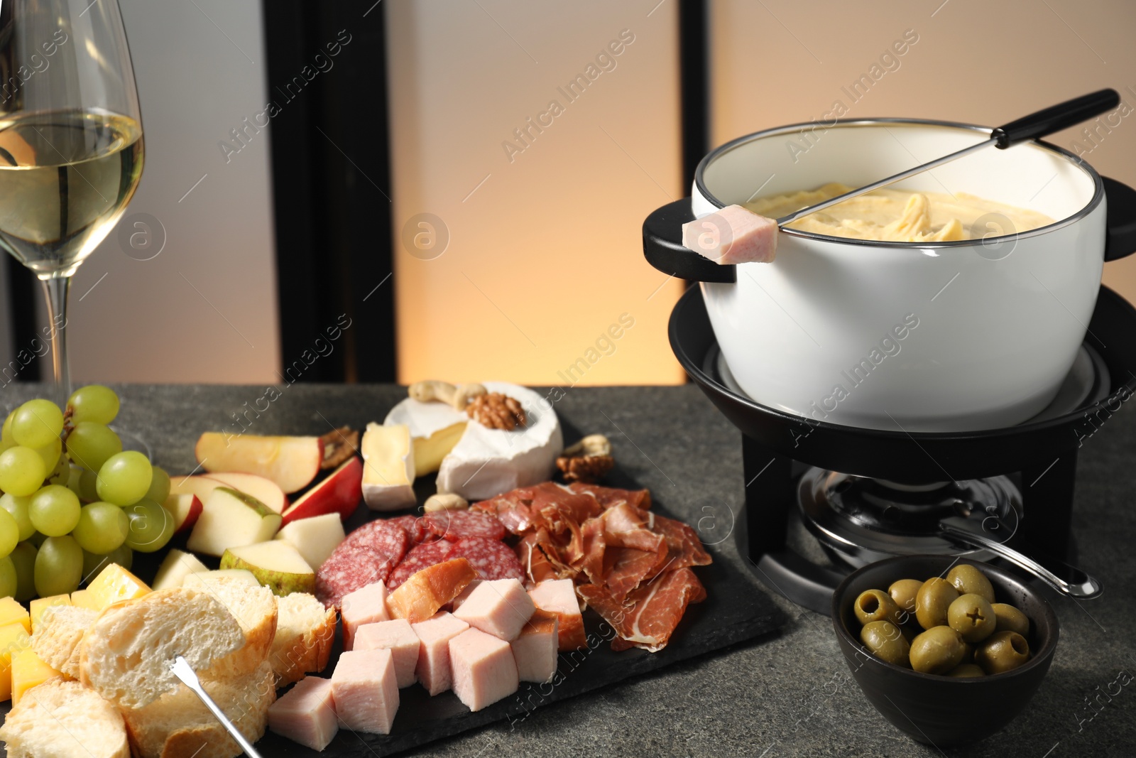 Photo of Fork with piece of ham, fondue pot with melted cheese, wine and snacks on grey table