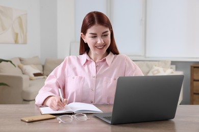 Photo of Happy woman with notebook using laptop at wooden table in room
