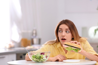 Emotional young woman eating sandwich instead of salad in kitchen. Healthy diet