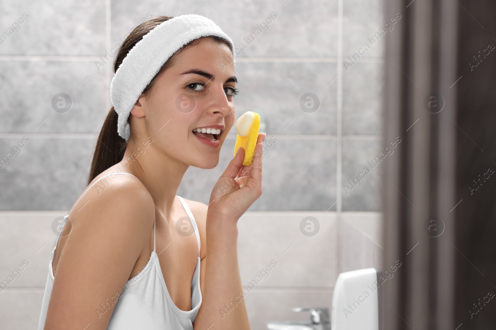 Photo of Young woman with headband washing her face using sponge in bathroom