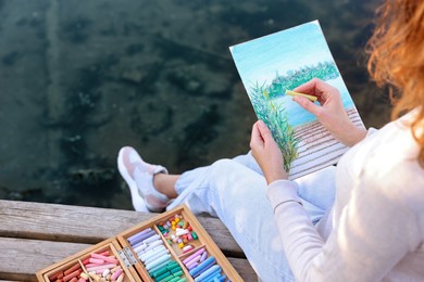 Photo of Woman drawing with soft pastels on wooden pier near river, closeup