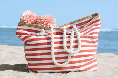 Stylish striped bag with slippers on sandy beach near sea
