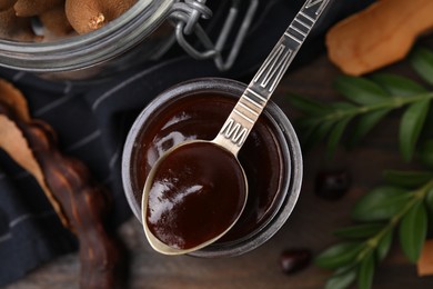 Photo of Jar of tasty tamarind sauce, spoon and fresh pods on wooden table, flat lay