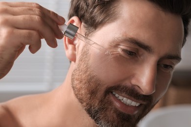 Smiling man applying cosmetic serum onto his face indoors, closeup