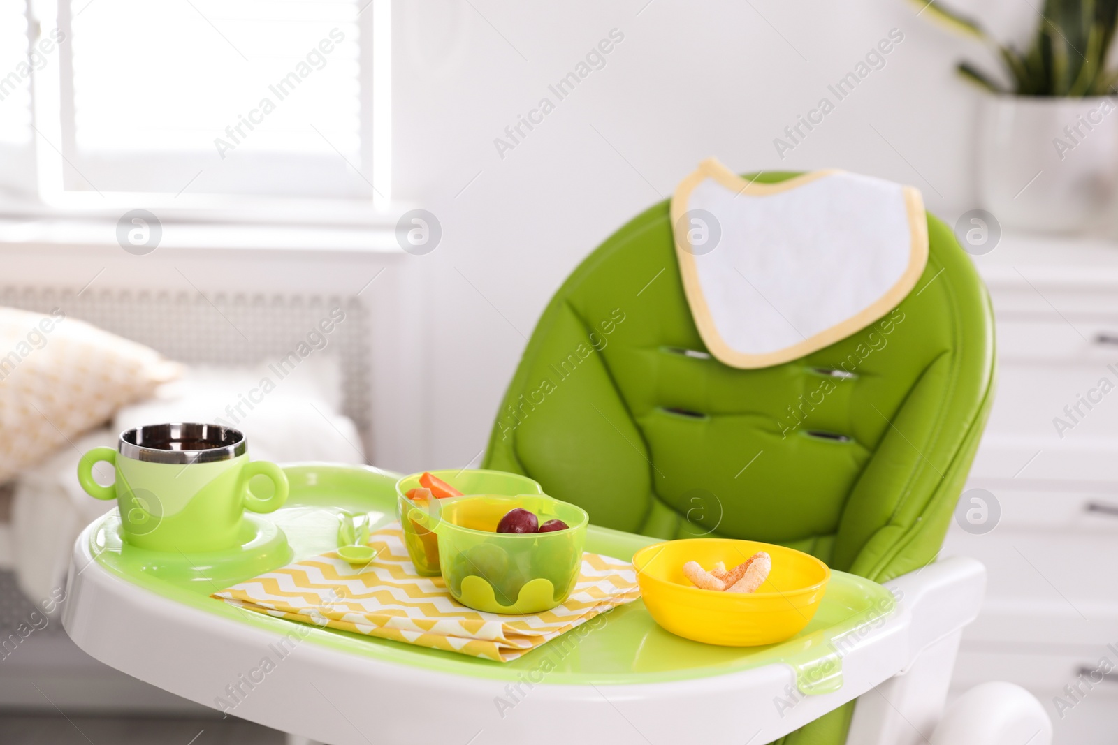 Photo of High chair with food in baby tableware on tray indoors