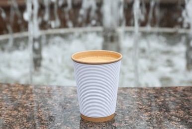 Photo of Paper cup of coffee near fountain outdoors. Takeaway drink