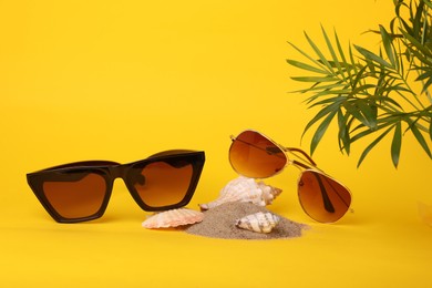 Stylish sunglasses, seashells, sand and palm leaves on yellow background