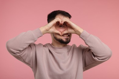 Photo of Man looking through folded in shape of heart hands on pink background