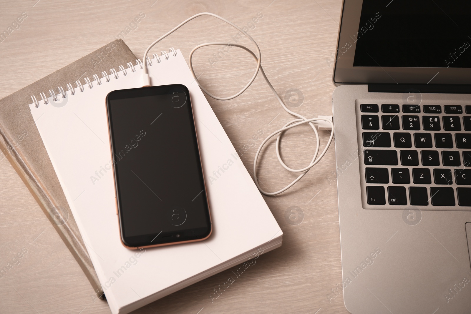 Photo of Smartphone connected with charge cable to laptop on wooden table, above view. Space for text