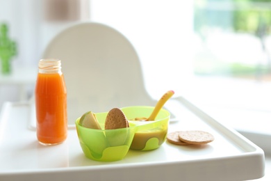 Bowls with delicious baby food and bottle of juice on highchair indoors