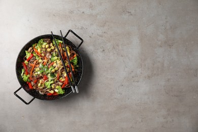 Photo of Stir-fry. Tasty noodles with meat in wok and chopsticks on grey textured table, top view. Space for text