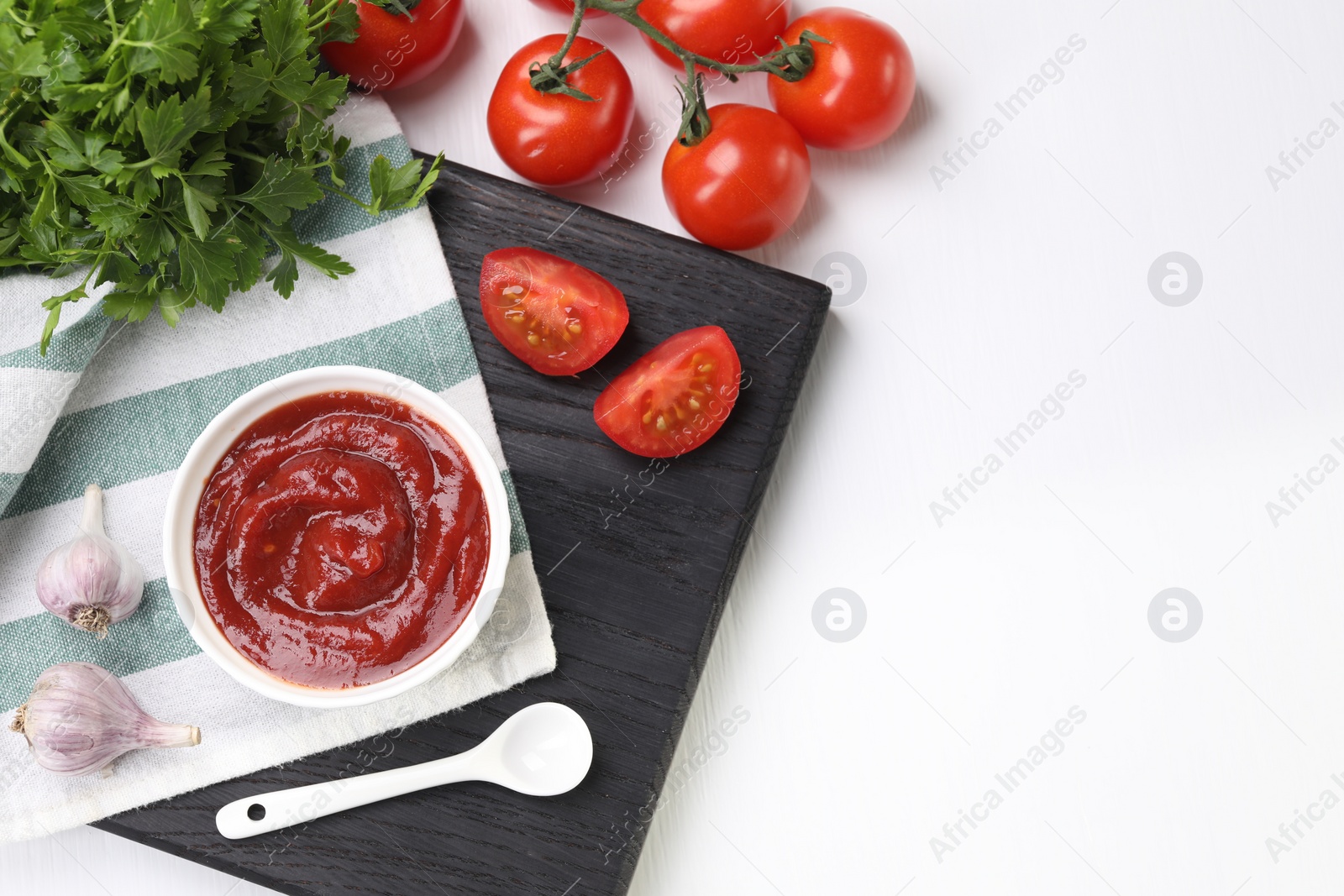 Photo of Organic ketchup in bowl, fresh tomatoes, parsley and garlic on white table, flat lay with space for text. Tomato sauce