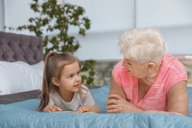 Cute girl and her grandmother reading book on bed at home