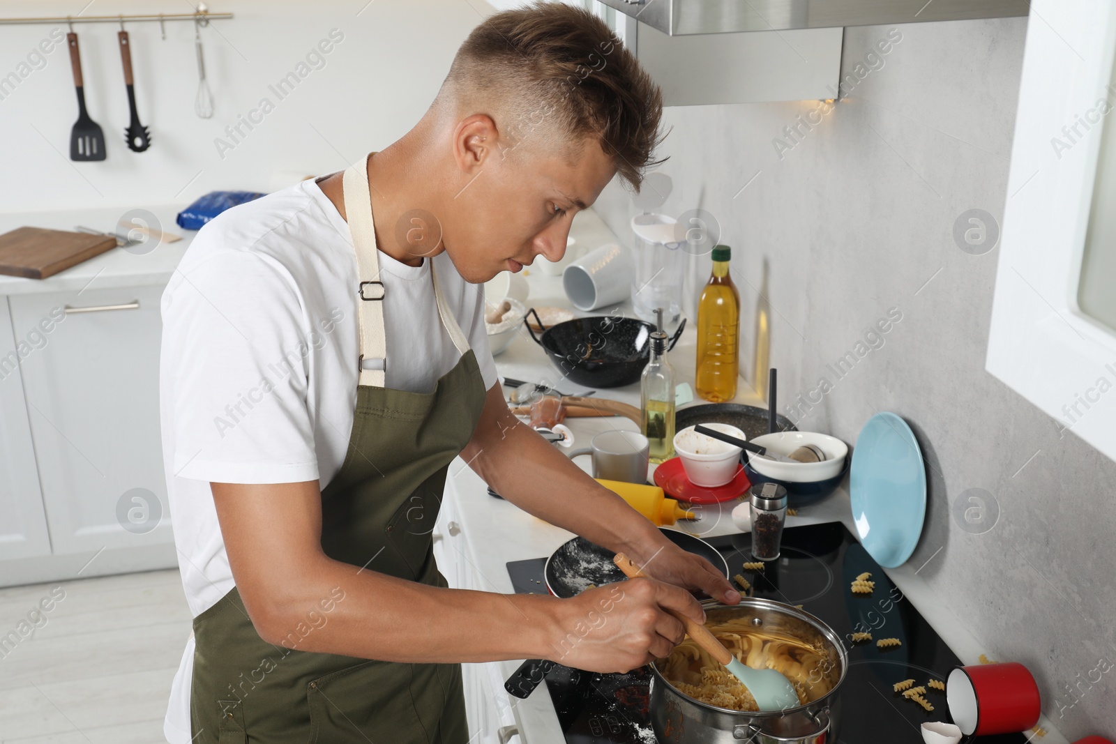 Photo of Man stirring pasta in saucepan in messy kitchen. Many dirty dishware and utensils on stove and countertop