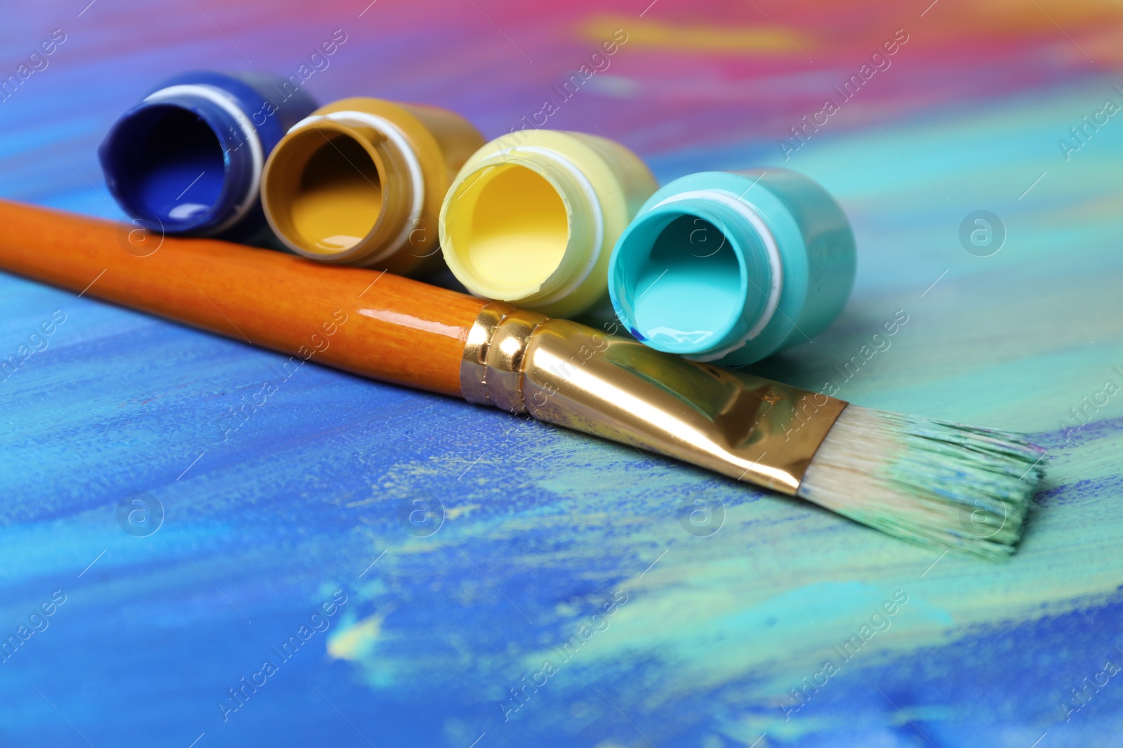 Photo of Jars with paints and brush on abstract picture, closeup