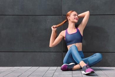 Photo of Beautiful woman in stylish gym clothes sitting near dark grey wall on street, space for text