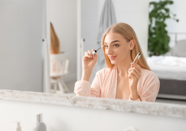 Photo of Young woman applying oil onto her eyelashes near mirror indoors