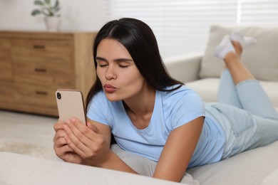 Photo of Young woman having video chat via smartphone and sending air kiss on sofa in living room