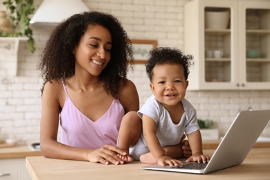 Photo of African-American woman and her baby with laptop in kitchen. Happiness of motherhood