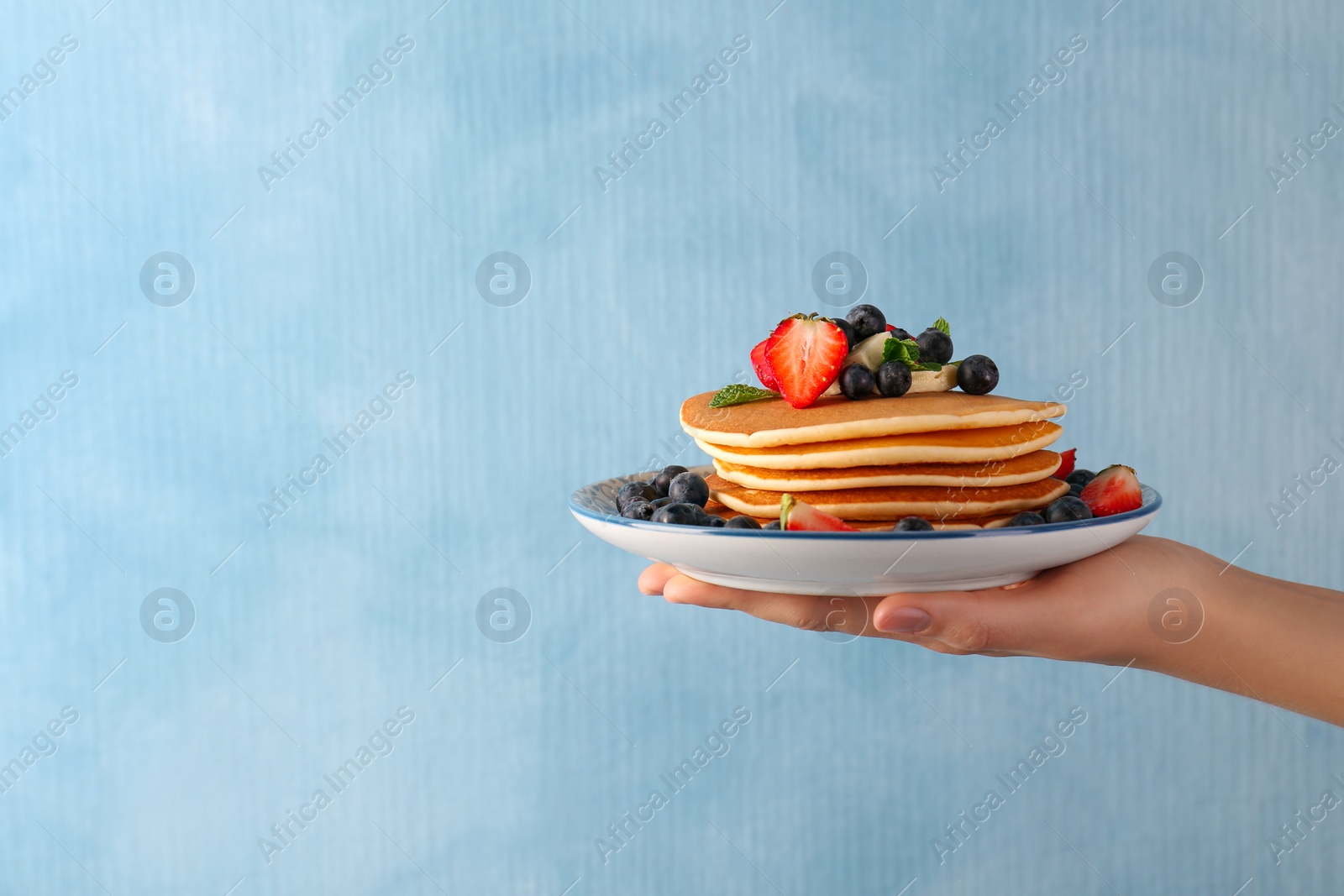Photo of Woman holding delicious pancakes with fresh berries and butter against light blue background, closeup. Space for text