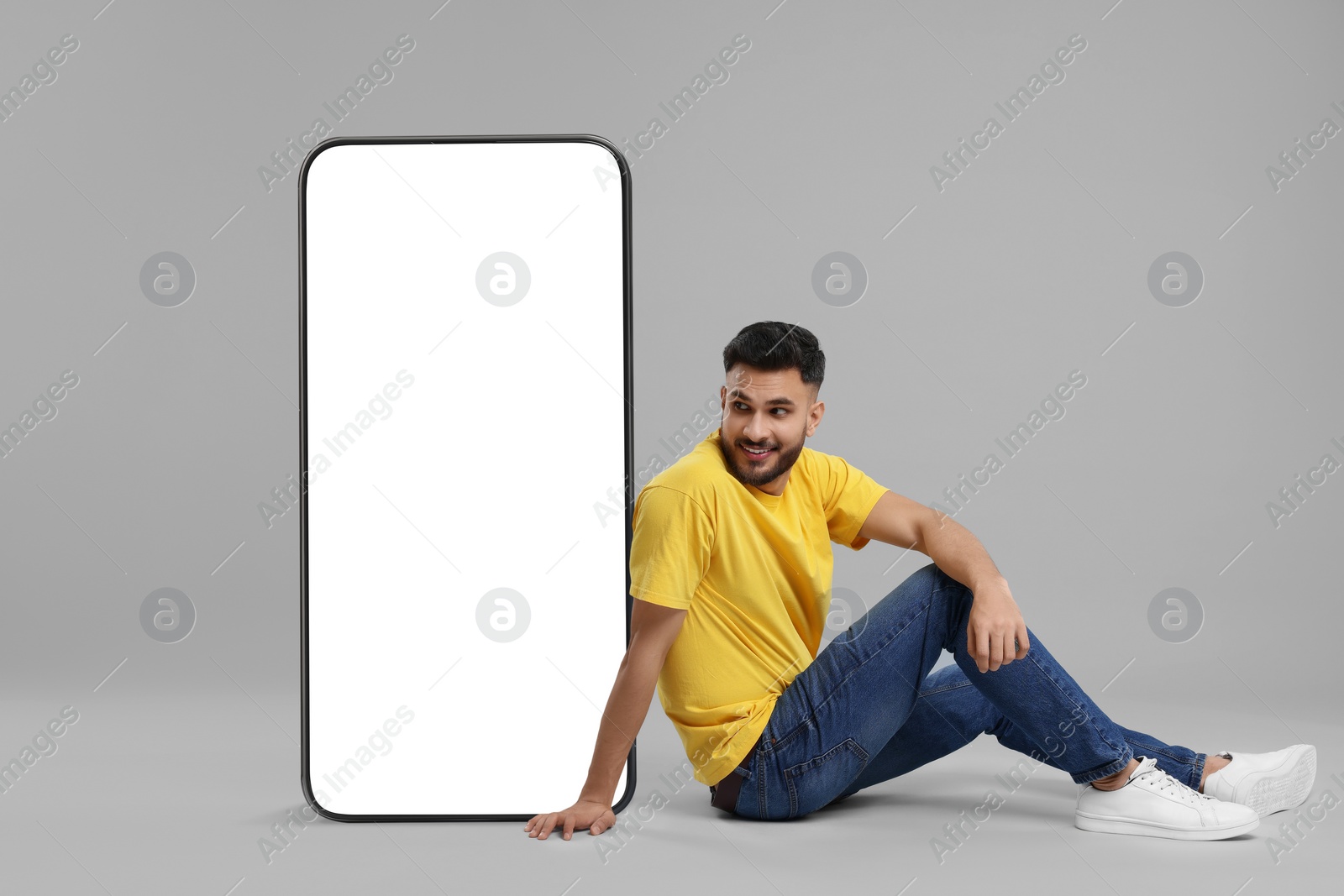 Image of Man sitting near huge mobile phone with empty screen on grey background. Mockup for design