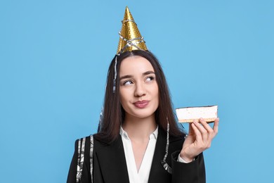 Photo of Woman in party hat with piece of tasty cake and streamers on light blue background