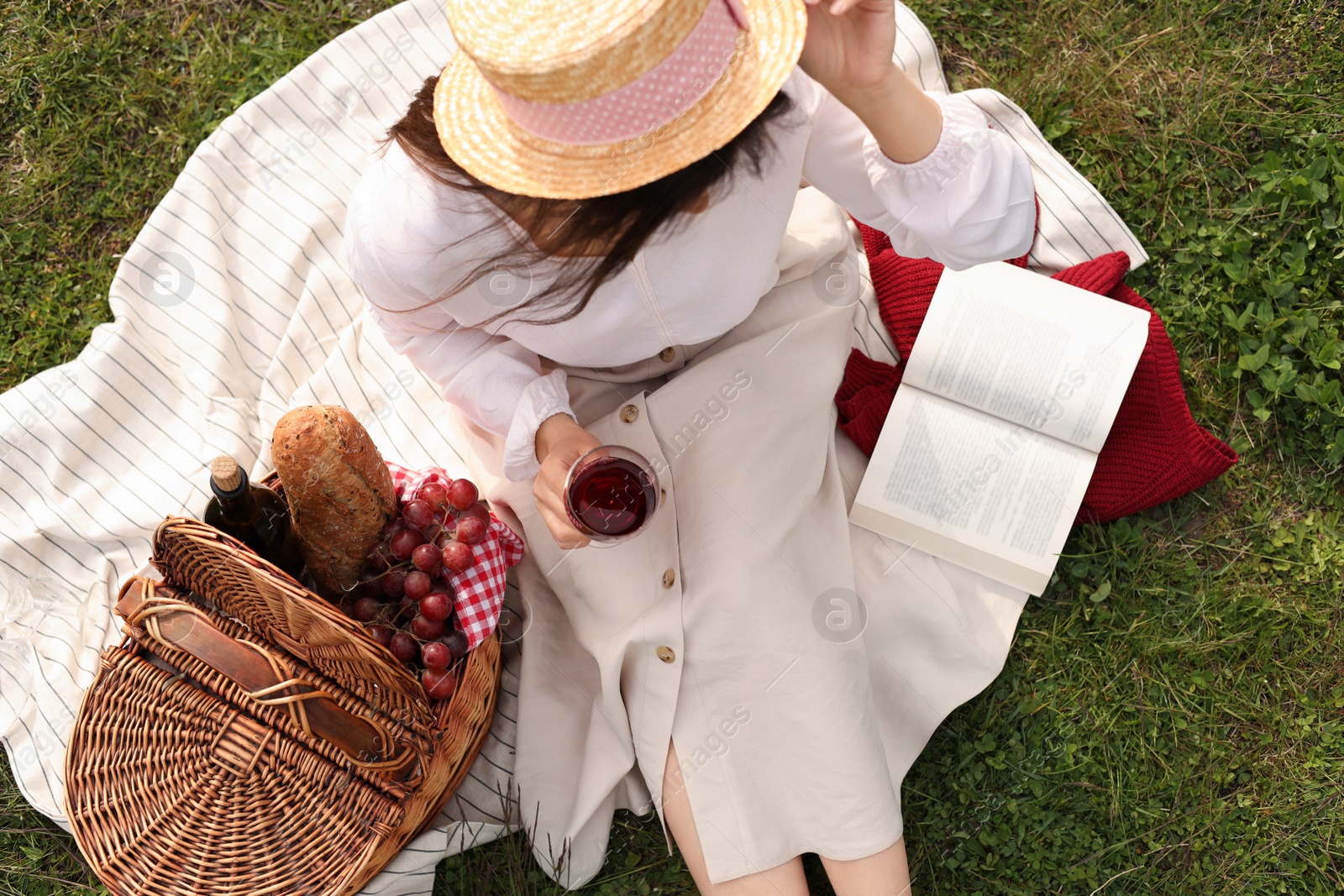 Photo of Woman with glass of wine, book and picnic basket on green grass, above view