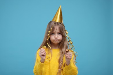 Upset little girl in party hat with confetti on light blue background