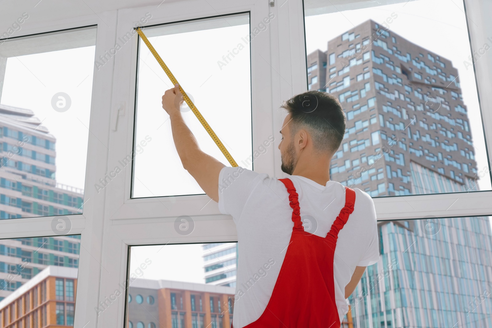 Photo of Worker in uniform measuring window with tape indoors, back view. Roller blinds installation