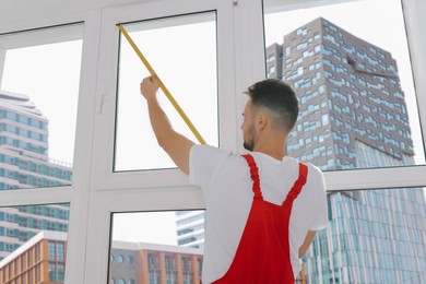 Photo of Worker in uniform measuring window with tape indoors, back view. Roller blinds installation
