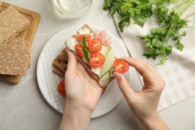 Photo of Woman eating fresh rye crispbread with cream cheese and vegetables at light grey marble table, top view