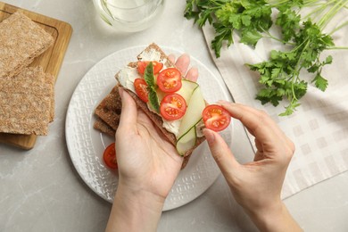 Woman eating fresh rye crispbread with cream cheese and vegetables at light grey marble table, top view