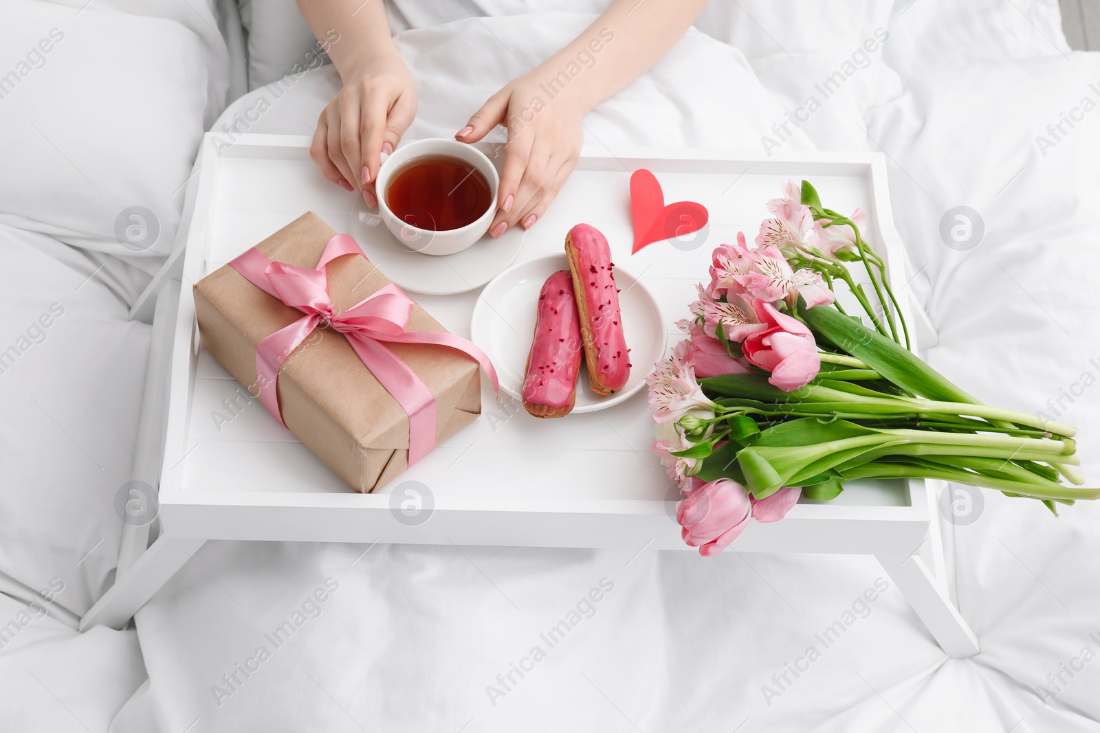 Photo of Tasty breakfast served in bed. Woman with tea, eclairs, gift box and flowers at home, closeup