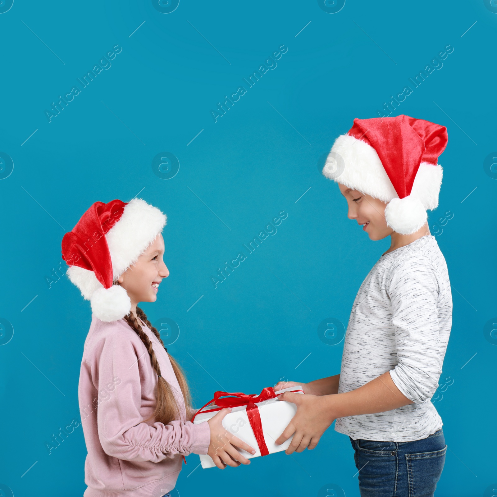 Image of Happy little children in Santa hats with Christmas gift on light blue background